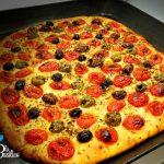 Focaccia barese appena sfornata con pomodorini tagliati a metà e olive verdi su teglia unta d'olio