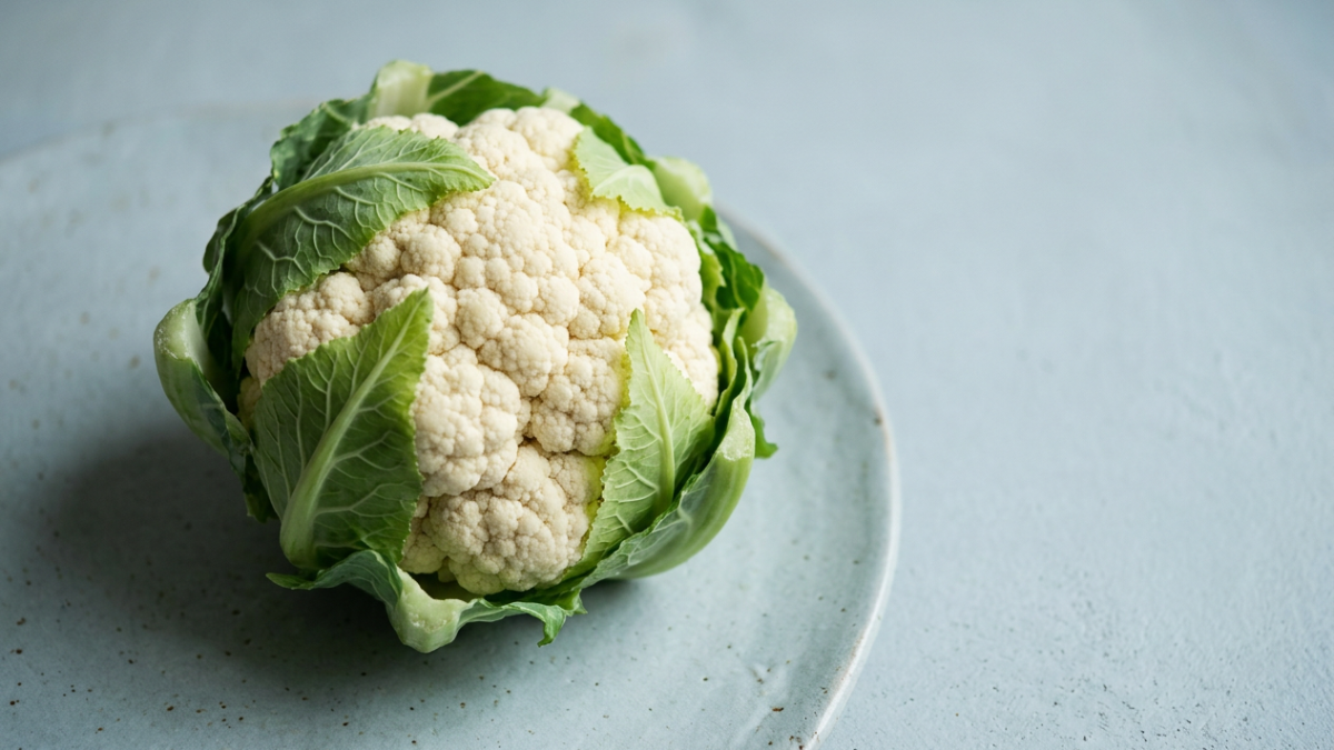 Un cavolfiore intero crudo con le foglie appoggiato su una ceramica azzurro-grigia.